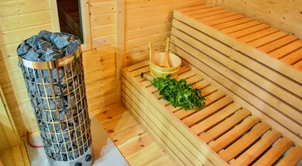 Interno di una sauna fai da te con stufa in pietra, panche in legno, secchiello e frusta di betulla pronti per un relax rigenerante.
