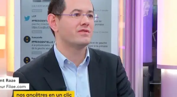 Un homme en costume, lunettes, parle. Le nom de famille gratuit "Filae.com" est visible, suggérant une recherche d'ancêtres.