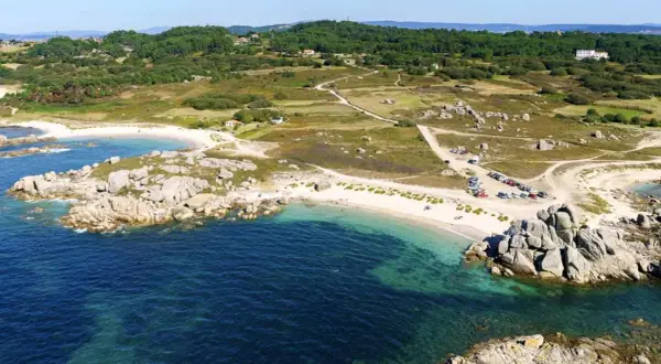 Vista aérea de una playa con arena blanca y aguas turquesas, rodeada de rocas y vegetación. Se ve un aparcamiento con coches cerca de la playa.