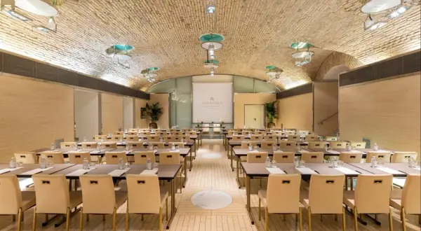 Sala riunioni con tavoli e sedie disposti per un evento all'Anantara Palazzo Naiadi Rome Hotel. Soffitto a volta in mattoni e illuminazione moderna.