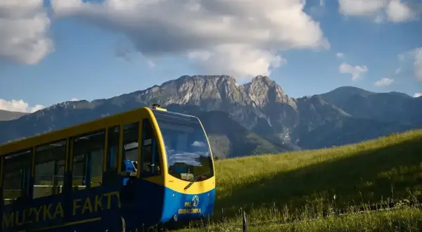 Żółto-niebieski wagonik PKL **wyciąg krzesełkowy na Gubałówkę** wjeżdża na zielone wzgórze, w tle majestatyczne Tatry.