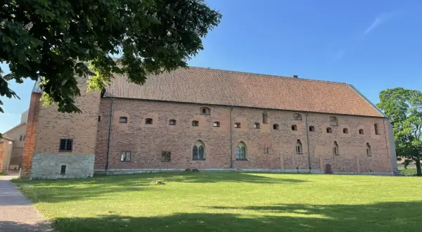 Imponerande tegelbyggnad med tegeltak, en del av Vadstena kloster, omgiven av grönskande gräsmatta och träd under en klarblå himmel.