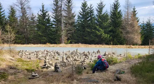 Osoba z plecakiem odpoczywa przy drodze, otoczona kamiennymi wieżami. W tle las i niebo. To miejsce może być częścią szlaku na jagodna szczyt.