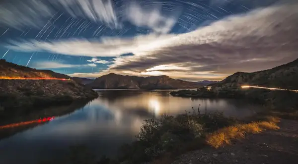 A surreal moon landscape with star trails arching over a calm lake and rugged mountains. Light trails from cars streak across the scene.