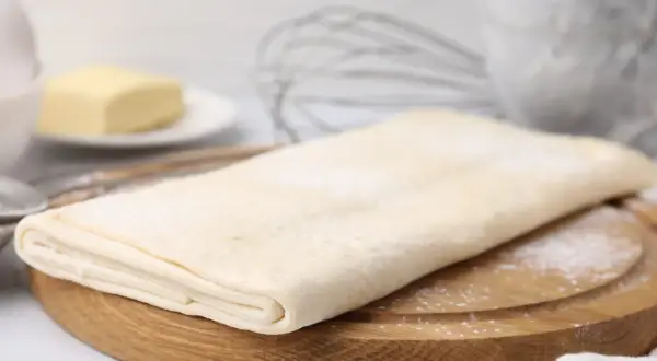Gotowe ciasto francuskie na desce, obok masło i trzepaczka. Sprawdź, jaka jest ciasto francuskie cena!