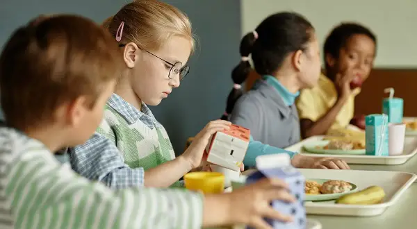 Kinder essen in der Mensa. Ein Mädchen mit Brille hält eine Milchpackung. Ein Teil des Tagesablaufs eines Schulbegleiters.