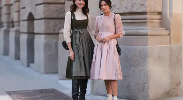 Zwei Frauen posieren vor einer Steinfassade. Eine trägt ein grünes Dirndl mit weißer Bluse und schwarze Stiefel.
