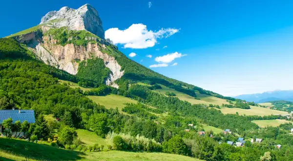 Vue panoramique d'une montagne imposante, le GR 965, dominant une vallée verdoyante parsemée de maisons. Le ciel bleu est parsemé de quelques nuages blancs.