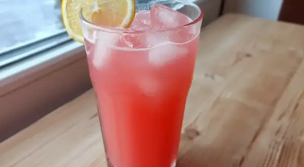 Ett glas rosa rabarberlemonad med is och en citronskiva, redo att avnjutas vid fönstret.