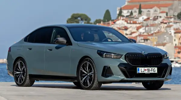 Szary sedan BMW 5 2025 z czarnym grillem i felgami, zaparkowany na tle malowniczego miasta nad morzem.