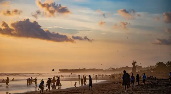 Zachód słońca na plaży Bali. Ludzie spacerują, grają w piłkę i puszczają latawce. W tle widać palmowy las.