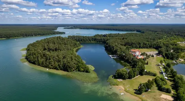 Malownicze jezioro Woświn otoczone lasem, z hotelem i polem kempingowym na brzegu. Błękitne niebo z białymi chmurami.