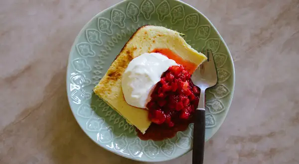 En bit ostkaka med keso, toppad med grädde och jordgubbar, på en mintgrön tallrik med en gaffel.