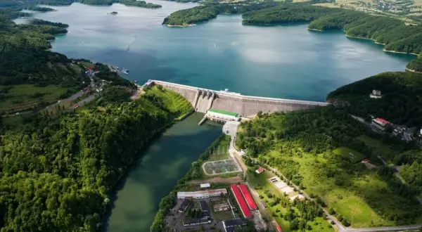 Ogromna zapora na sanie spiętrza turkusową wodę jeziora otoczonego zielonymi wzgórzami. Widać budynki elektrowni i infrastrukturę.