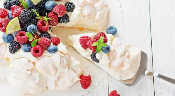 Beza Pavlova która zawsze wychodzi, udekorowana świeżymi malinami, borówkami, jeżynami i listkami mięty. Kawałek ciasta jest już nakładany.