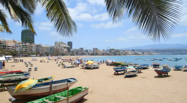 Solig strand i Las Palmas med palmer, båtar på sanden och en livlig stad i bakgrunden.