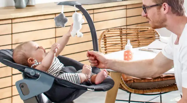 Ein Vater spielt mit seinem Baby in einem Hochstuhl. Ab wann Babys im Hochstuhl sitzen, hängt von ihrer Entwicklung ab.