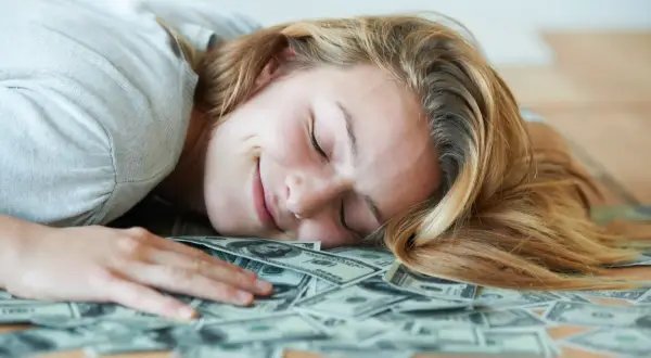 Uśmiechnięta kobieta śpi na stosie banknotów dolarowych. Marzy o tym, czy polskie gwiazdy rzeczywiście biorą udział w międzynarodowych kumulacjach.