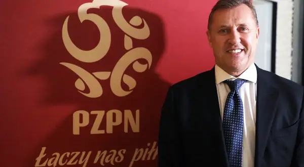 Uśmiechnięty prezes PZPN w garniturze stoi obok czerwonego banera z logo związku i hasłem "Łączy nas piłka".