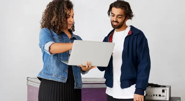 A woman and man review a laptop, discussing music artist management. They stand near stage equipment.