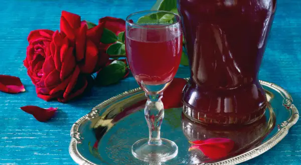 Aromatic nalewka z płatków róży w kieliszku i karafce, obok czerwona róża i płatki na srebrnej tacy.
