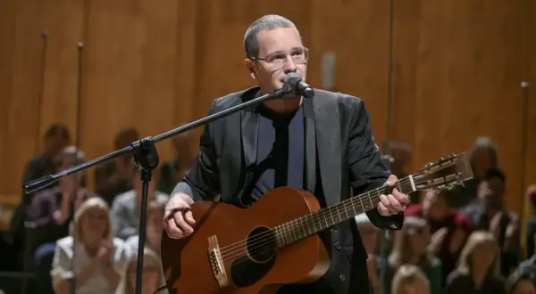 Artysta z gitarą i mikrofonem na scenie, przygotowuje się do koncertu w ramach **Męskie Granie 2026**. Publiczność w tle czeka na występ.