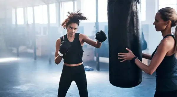Woman training boxing for fitness at home, punching a heavy bag while a trainer holds it steady.