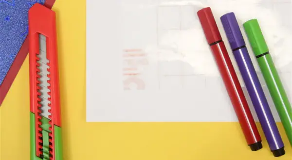 Un cutter rouge et trois feutres (rouge, violet, vert) sont prêts pour un projet créatif. Une **lettre noire** est visible sur le papier de fond.