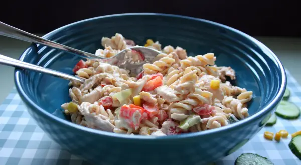 En stor blå skål fylld med krämig pastasallad med kyckling, tomat och majs. Två gafflar ligger ovanpå.