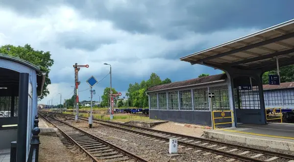 Tory kolejowe na stacji Starogard Gdański pod pochmurnym niebem. Widać peron, budynek poczekalni i semafory.