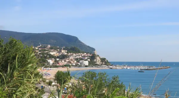 Vista panoramica della Riviera del Conero con un borgo arroccato su una collina, spiaggia attrezzata e un porto turistico.