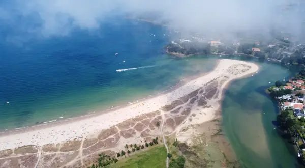 Vista aérea de la playa de Miño, con arena dorada, mar azul y verde, y casas costeras.
