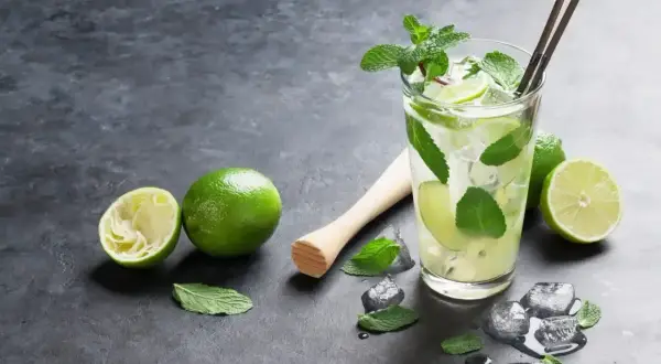 Orzeźwiający mohito smak: szklanka z lodem, limonkami i miętą, obok wyciśnięta limonka i tłuczek.