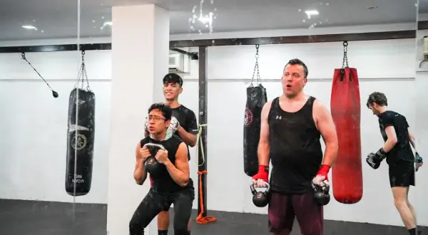 Men engage in boxing weight training, performing kettlebell squats and other exercises in a gym with punching bags.