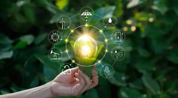 Dłoń trzyma żarówkę z zielonymi liśćmi, otoczoną ikonami symbolizującymi różne źródła energii, w tym OZE.