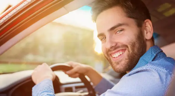 Un homme souriant au volant de sa voiture, prêt pour sa previsite contrôle technique. Le soleil brille, annonçant une journée sereine.