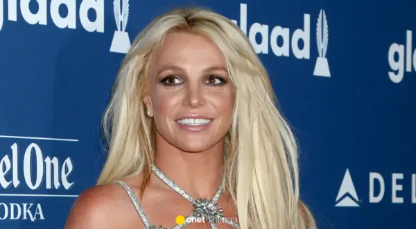 Uśmiechnięta Britney Spears, której wiek nie jest widoczny na zdjęciu, w błyszczącej kreacji na tle niebieskiej ścianki z logo GLAAD.