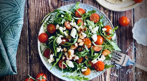 Frischer Rucola Salat mit Kirschtomaten, Feta und Croutons auf rustikalem Holztisch.
