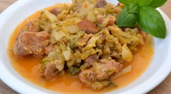 Pyszny bigos z młodej kapusty z kawałkami mięsa, podany na białym talerzu, ozdobiony gałązką bazylii.