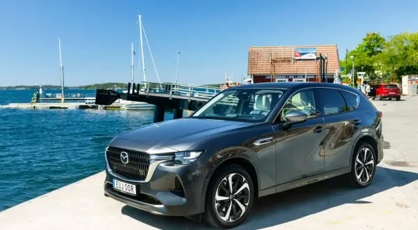 Mörkgrå Mazda CX-60 laddhybrid parkerad vid en brygga med båtar och en röd bil i bakgrunden. En solig dag för ett Mazda CX-60 laddhybrid test.