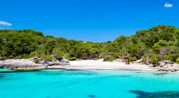 Aguas turquesas y arena blanca en la cala turqueta menorca, rodeada de pinos y rocas. Un paraíso balear.