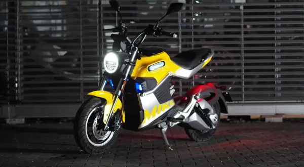 Żółty motocykl elektryczny z okrągłym reflektorem, stojący na kostce brukowej przed metalową fasadą.