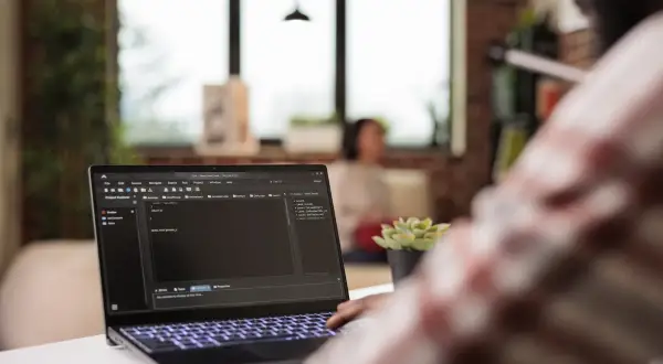Programista pracuje nad kodem na laptopie, gdzie widać plik hosts. W tle siedzi osoba.