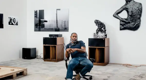 A man sits in an office chair in a gallery space, surrounded by art and audio equipment, a scene that feels like a snapshot of Arthur Jafa's creative process.