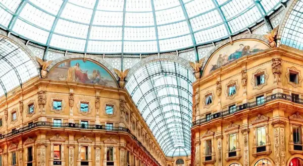 Inside Milan's Galleria Vittorio Emanuele II, a stunning glass dome arches over ornate buildings adorned with frescoes, reminiscent of grand Milan art galleries.