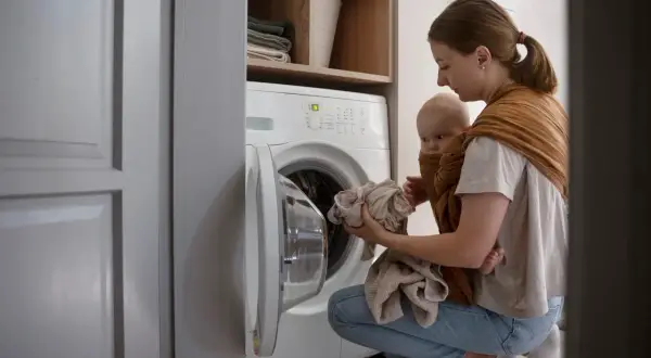 Une mère porte son bébé en écharpe tout en chargeant le linge dans le lave-linge. Une astuce de grand-mère pour éviter la mauvaise odeur du lave-linge.