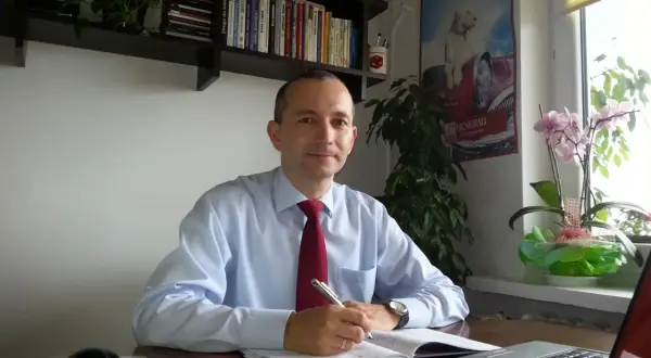 Mężczyzna w koszuli i krawacie siedzi przy biurku, pisząc w notatniku.