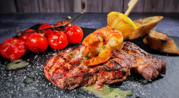 Perfektes Surf and Turf Rezept: saftiges Steak mit gegrillten Garnelen, Kirschtomaten und Zitronenspalte auf schwarzem Stein.