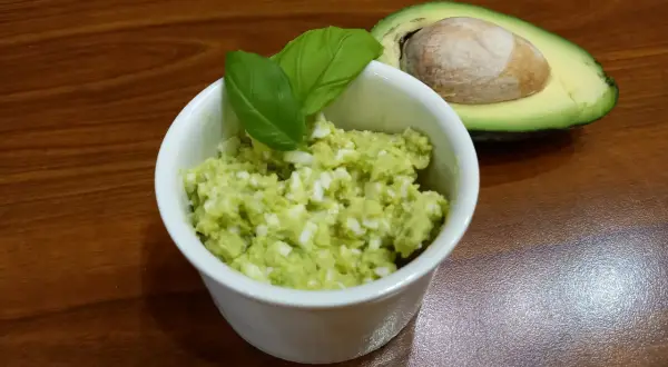 Pyszna pasta z awokado i jajka, udekorowana listkami bazylii, obok połówka awokado z pestką.