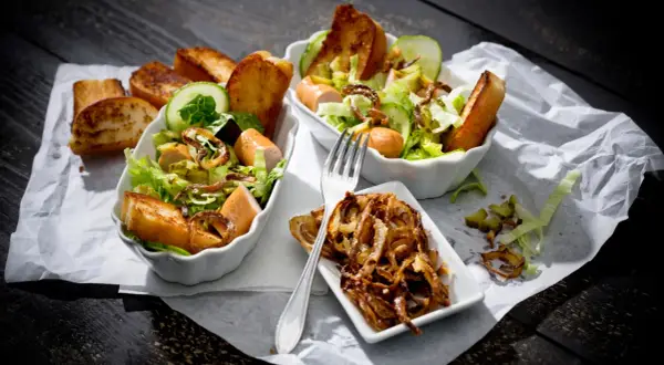 Zwei Schüsseln mit Hot Dog Salat, garniert mit Gurkenscheiben, geröstetem Brot und knusprigen Zwiebeln.